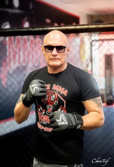 Portrait d'un homme chauve avec gants de MMA posant devant une cage de combat à Perpignan 66.