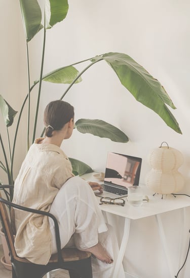 A woman in beige shirt and white trouser sitting at a desk looking into the laptop. Calm scene