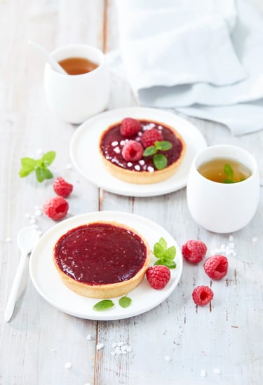 deux tartelettes aux framboises sur un fond blanc cérusé avec quelques framboises éparpillé