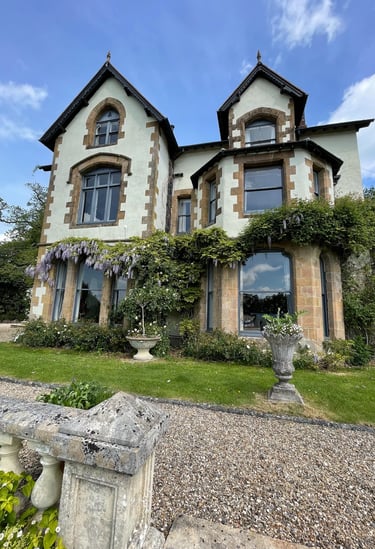A historic country house featuring gothic windows, stone architecture, and purple wisteria climbing the walls.