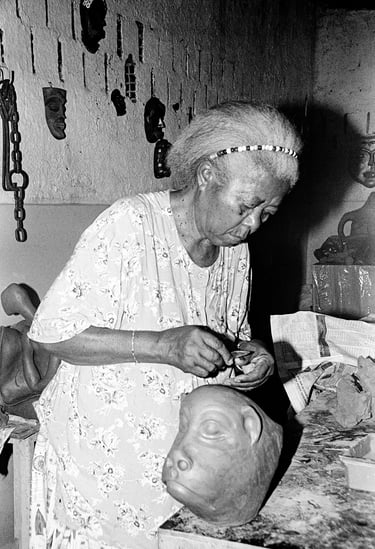 Ana of Carrancas sculpting a clay head in her studio with traditional masks displayed.