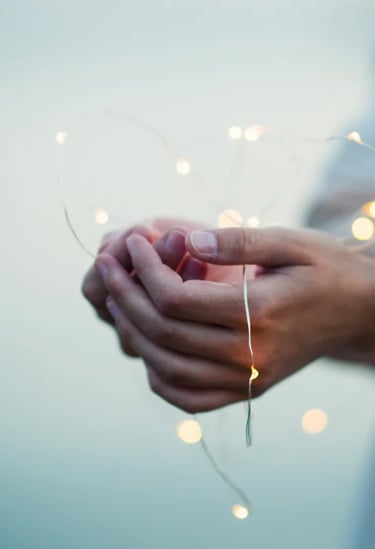Cupped hands holding warm glowing fairy lights in the shape of a heart against a soft blue background.