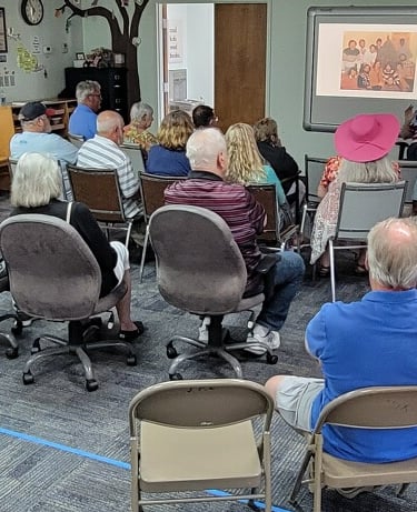 A group of senior citizens attending a community presentation with a projector screen in a meeting room.