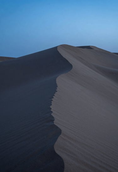 Clean, modern photography of desert sand dunes at twilight, focusing on the elegant curves and sharp shadows, deep blue and slate grey tones, minimalistic and serene aesthetic.