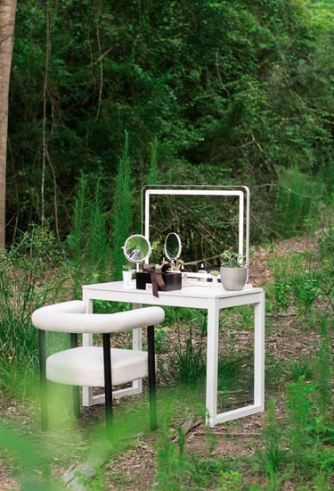A modern white vanity table with a lighted mirror and armchair set outdoors in a lush green forest.