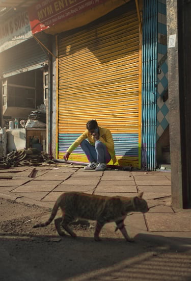Man squatting outside a closed shop as a cat walks past in Old Delhi