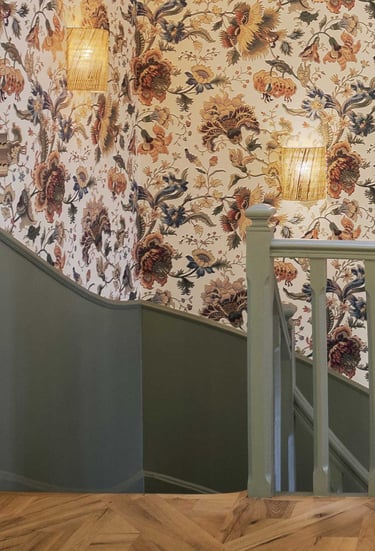 Vintage floral wallpaper lines a staircase with sage green wood paneling and woven wall sconces.