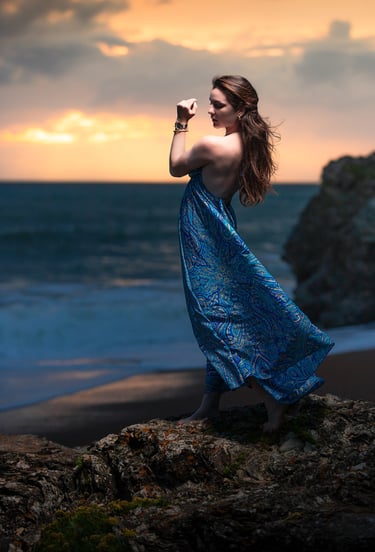 Portrait créatif d’une femme en robe bleue posant sur des rochers face à la mer au coucher du soleil