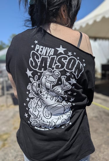 Person wearing black off-shoulder t-shirt with white cartoon frog smoking, flames, stars, and "PEÑA SALSON" text on the back
