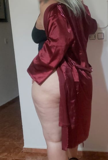 Escort madura colombiana en bata roja de satén, de rodillas sobre el sofá, mostrando sus piernas provocativas.