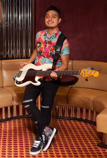 Bassist Ian Yoshio leaning against a coffee table, wearing a colorful shirt, with his white Fender bass guitar.