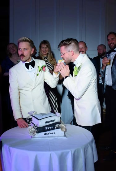 Grooms in white tuxedos eating cake at their wedding reception with guests watching.