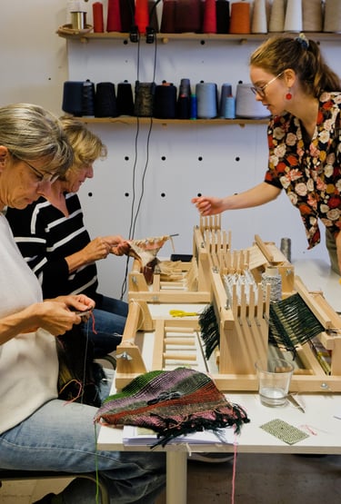 Weaving workshop instructor assisting students with small table looms and colorful yarn.