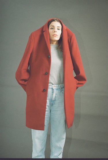 A woman wearing a large red wool coat over her head with light blue jeans against a grey background.