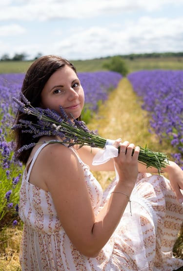Woman sitting in a lavender field, wearing a cream flowing dress and holding a bunch of lavender