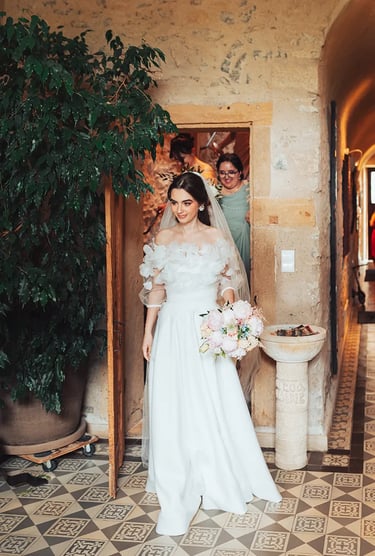 Anaïs, prête pour son mariage, sortant du gîte en robe de mariée avec son bouquet.