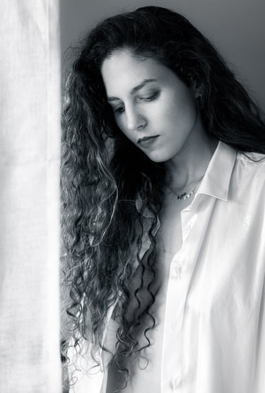 Portrait fine art en noir et blanc d’une femme aux cheveux longs, posant près d’une fenêtre