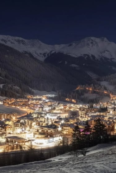 A panoramic, nighttime view of an Alpine ski resort town nestled in a valley, with glowing lights vi