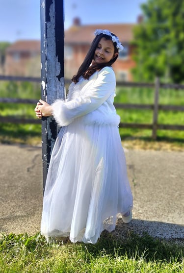 Close-up of a young girl with long brown hair, wearing a white dress and a floral white headband, smiling outdoors on a sunny