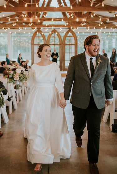 Smiling bride and groom holding hands walking down the aisle in a rustic barn wedding venue.