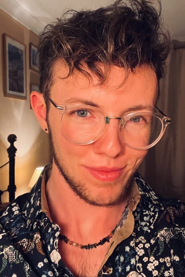White trans masculine person with blue eyes, glasses, light brown curls and facial hair, smiling