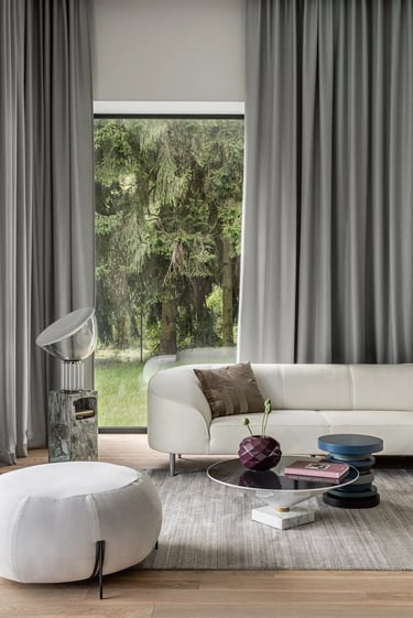 Aranżacja jasnego salonu, biała skórzana sofa, designerskie stoliki kawowe i szare zasłony.
