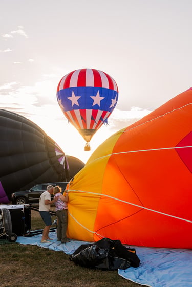 A vibrant patriotic hot air balloon takes flight as crews inflate more balloons during a sunrise festival.