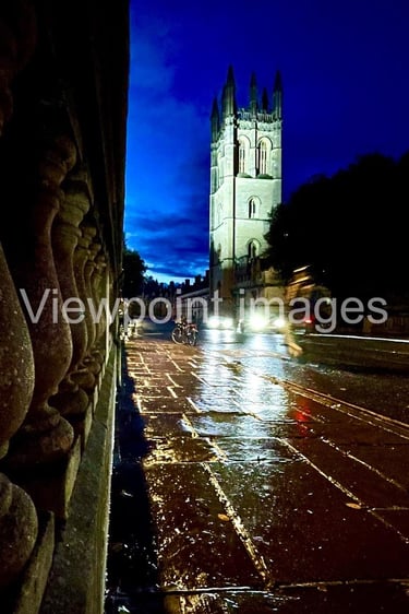 Magdalen College Tower at night viewed from a rainy Oxford street with reflections on wet pavement.