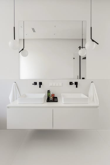 Minimalistyczna biała łazienka, szafka z dwema umywalkami, duże lustro i czarna armatura podtynkowa.