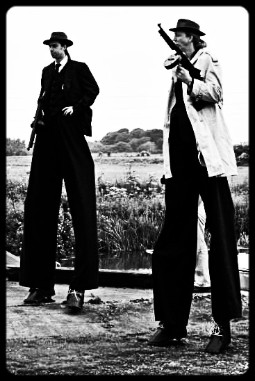 Black and white photo of two men on tall stilts dressed as gangsters holding prop tommy guns.