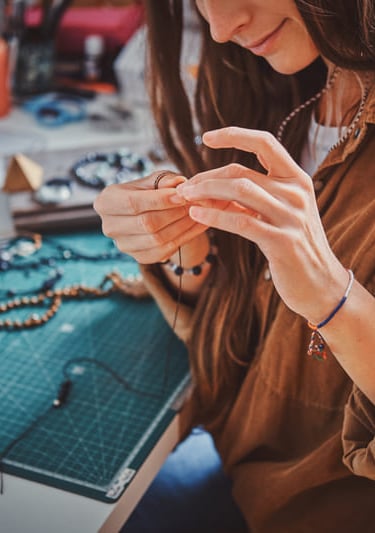 mujer sosteniendo joyeria artesanal frente a mesa de trabajo llena de cuentas e hilo