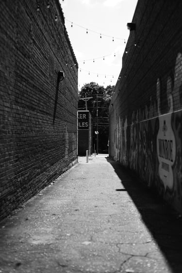 Black and white photo of a narrow brick urban alleyway with hanging lights and wall murals.