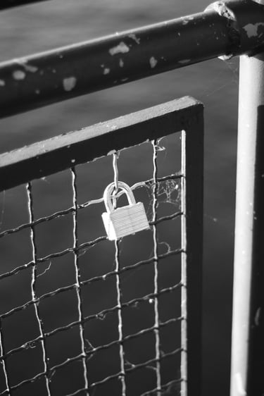 Black and white photo of a metal love lock attached to a wire mesh bridge railing.