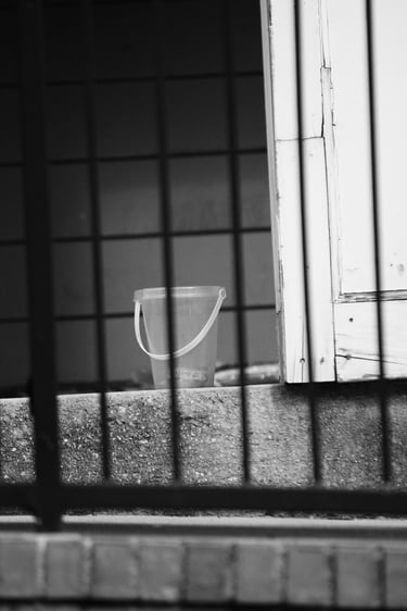 Black and white photo of a plastic bucket sitting on concrete steps behind a metal security fence.