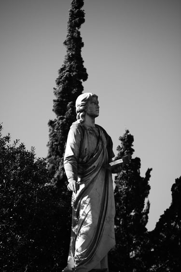 Black and white marble religious statue of a saint standing before tall cypress trees.