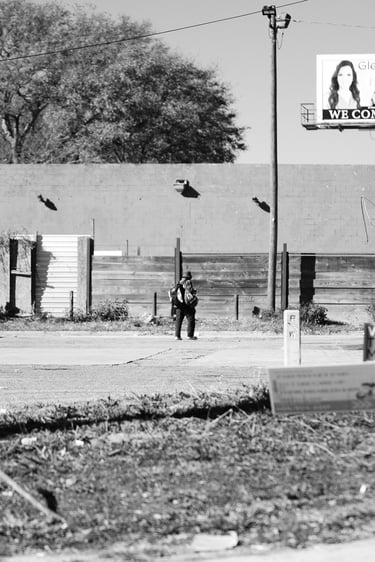 Black and white street photography of a person walking past urban billboards and a parking lot.