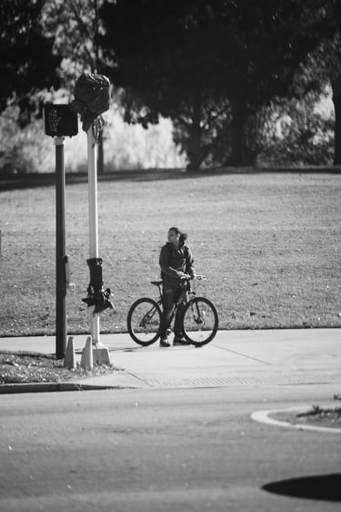 A cyclist stands with a mountain bike on a sunny paved path near a grassy park in black and white.
