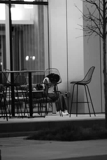 A person resting their head at an outdoor cafe table at night in a black and white street photograph.