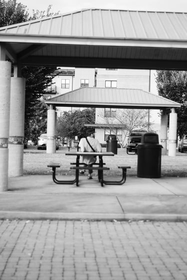 Black and white photo of a person sitting at a park picnic table under a metal pavilion.