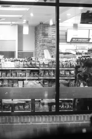 Black and white view of a person sitting in a convenience store near snacks and a self-checkout kiosk.