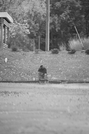Black and white street photography of a man sitting alone on a curb near a Wendy's restaurant.
