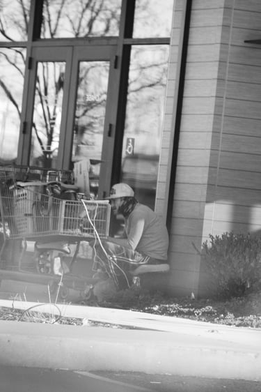 Black and white photo of a person sitting by a shopping cart filled with belongings outside a building.