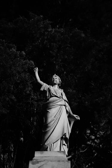 Black and white photo of a vintage stone angel statue in a cemetery surrounded by dark trees.