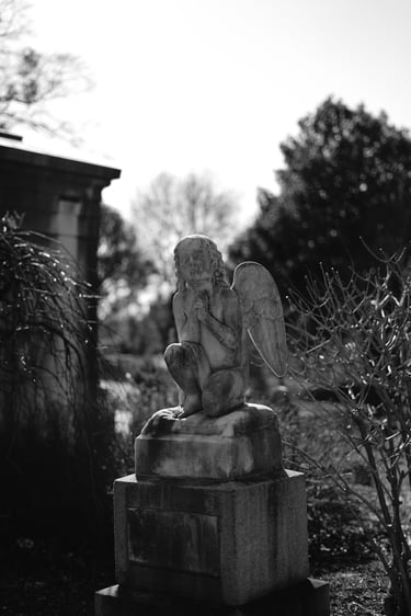 Black and white photo of a weathered stone angel statue praying on a grave in a peaceful cemetery.