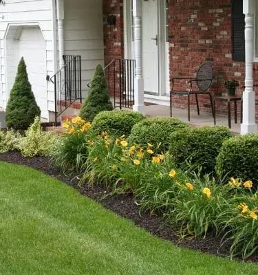 Beautifully landscaped front yard with vibrant green lawn, flowering plants, neatly trimmed bushes, and a brick house in the