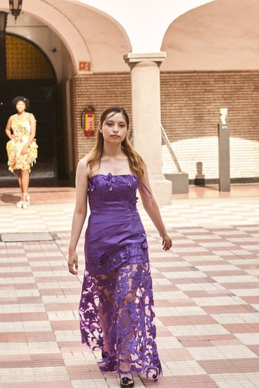 A model walks a runway in a purple strapless gown with a floral lace skirt during a fashion show.