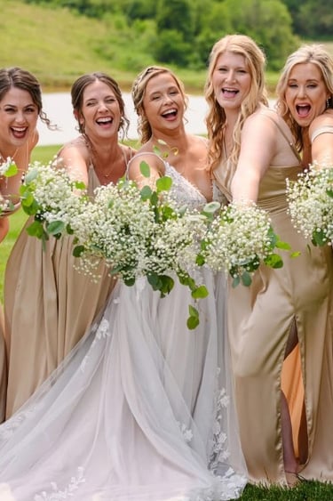 Bride surrounded by bridesmaids in gold dresses holding white bouquets, smiling outdoors, featuring wedding hair and makeup.