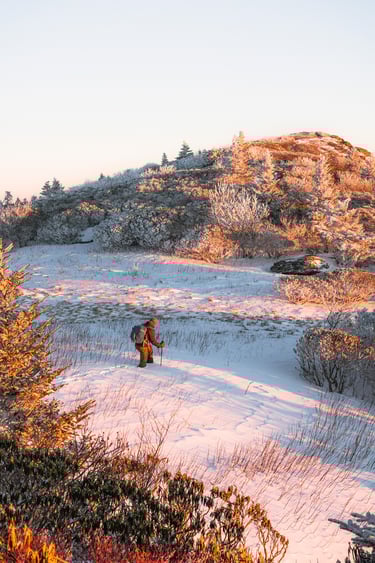 A hiker with trekking poles walks through deep snow on a mountain trail at sunrise.