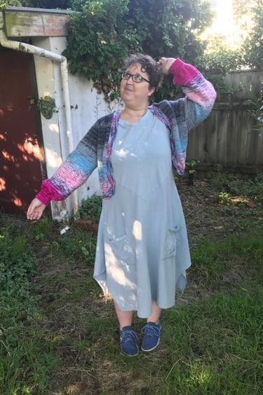 Photograph of Sarah Overdale posing in her garden,  wearing a dress and colourful knitwear