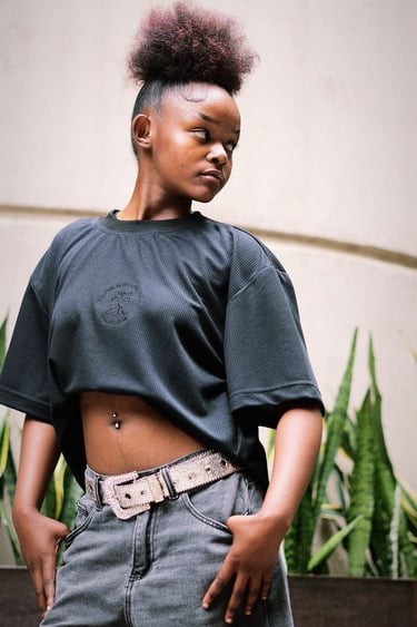 Young woman with a high puff hairstyle wearing a black crop top and grey jeans with a decorative belt.